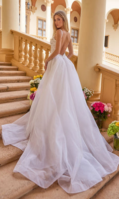 A woman wearing the Ava Presley 47224 long formal dress with a flowing tulle overskirt stands on stone stairs among floral arrangements in an outdoor architectural setting with columns and arches.