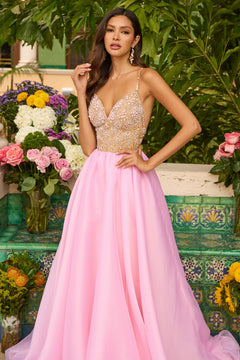 A woman stands outdoors among flowers and greenery, wearing the Ava Presley 47243 long prom dress featuring a beaded bodice and delicate spaghetti straps in light pink.