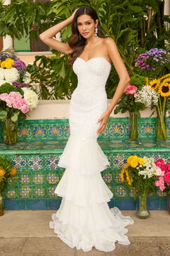 A woman in the ivory Ava Presley 47251 long prom dress, featuring a strapless mermaid silhouette and tiered ruffle layers, stands before colorful tiles and vibrant floral arrangements.