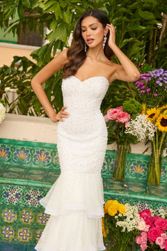 A woman poses outdoors by a colorful tiled wall and vibrant flowers, wearing the Ava Presley 47251 long strapless mermaid prom dress with pearl embellishments.