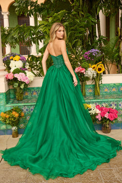 A woman stands outside among lush plants and vibrant flowers, her back to the camera, wearing the Long Prom Dress: Ava Presley 47255 featuring a strapless design and an embellished bodice.