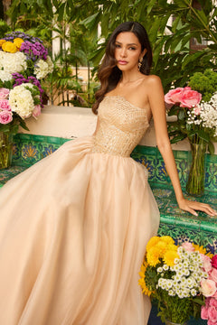 A woman in the Ava Presley 47255 Long Prom Dress, a strapless ball gown with an embellished bodice, sits on a tiled bench amid colorful flowers and lush greenery.