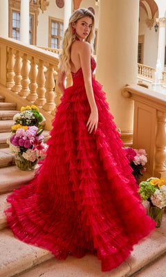 A woman wearing the Ava Presley 47258 long prom dress, a strapless red layered ruffled ball gown, stands on a staircase adorned with flowers in an elegant building with columns and arches.