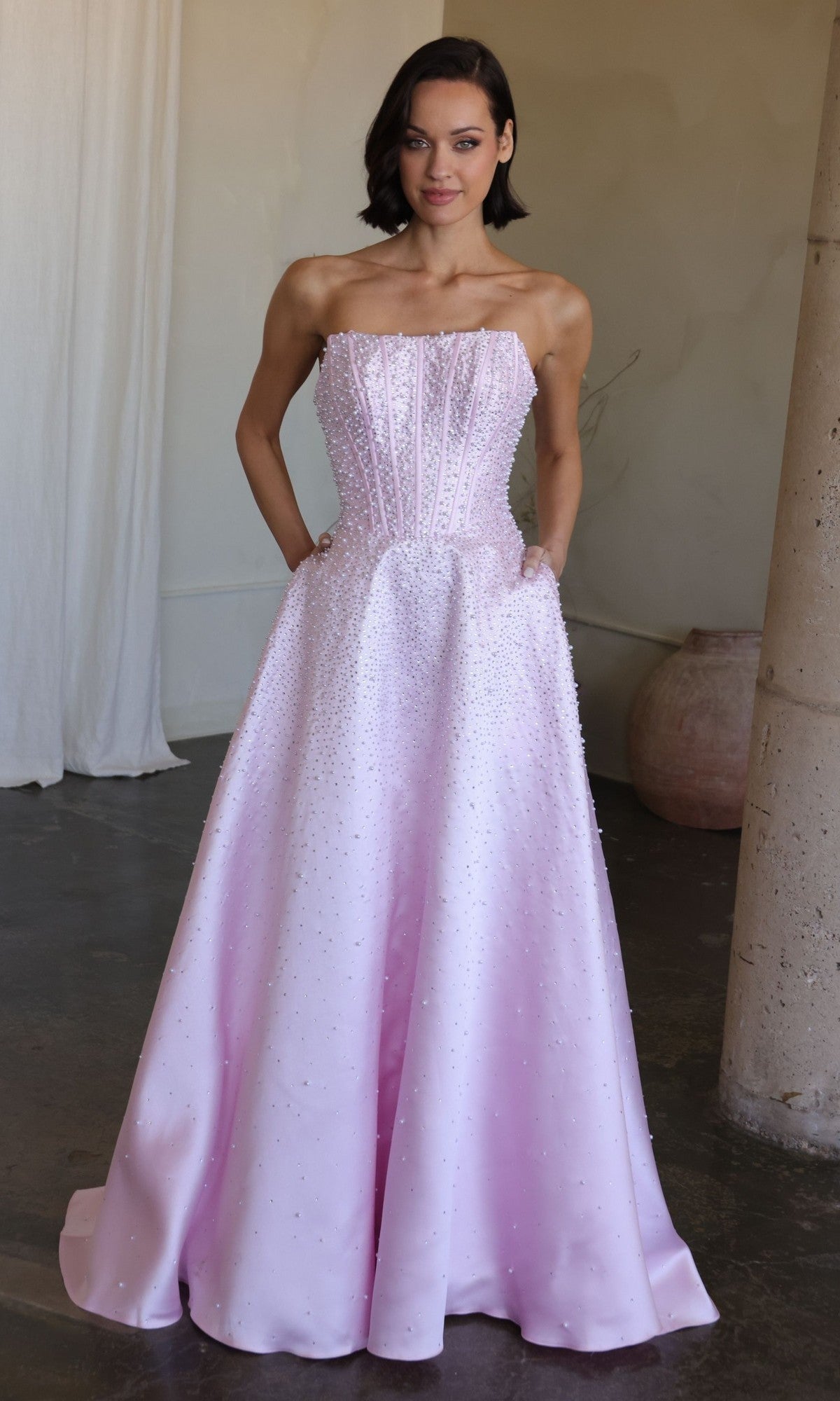 A woman stands indoors, hands in pockets, wearing the Ava Presley 47283 long prom dress featuring a light pink strapless design with a fitted, beaded corset bodice.