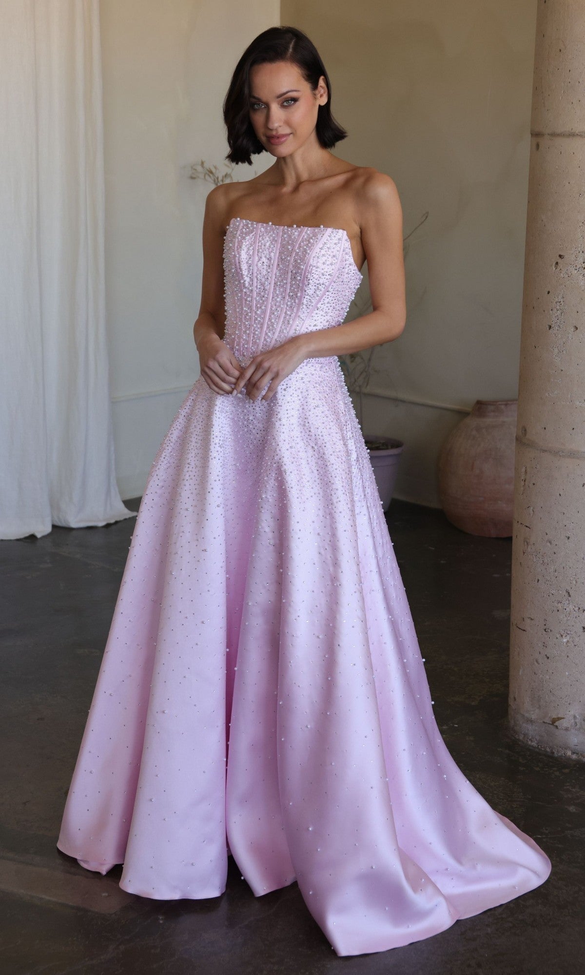 A woman indoors wears the Ava Presley 47283 long prom dress in blush, featuring a strapless corset bodice and floor-length skirt adorned with delicate bead details.