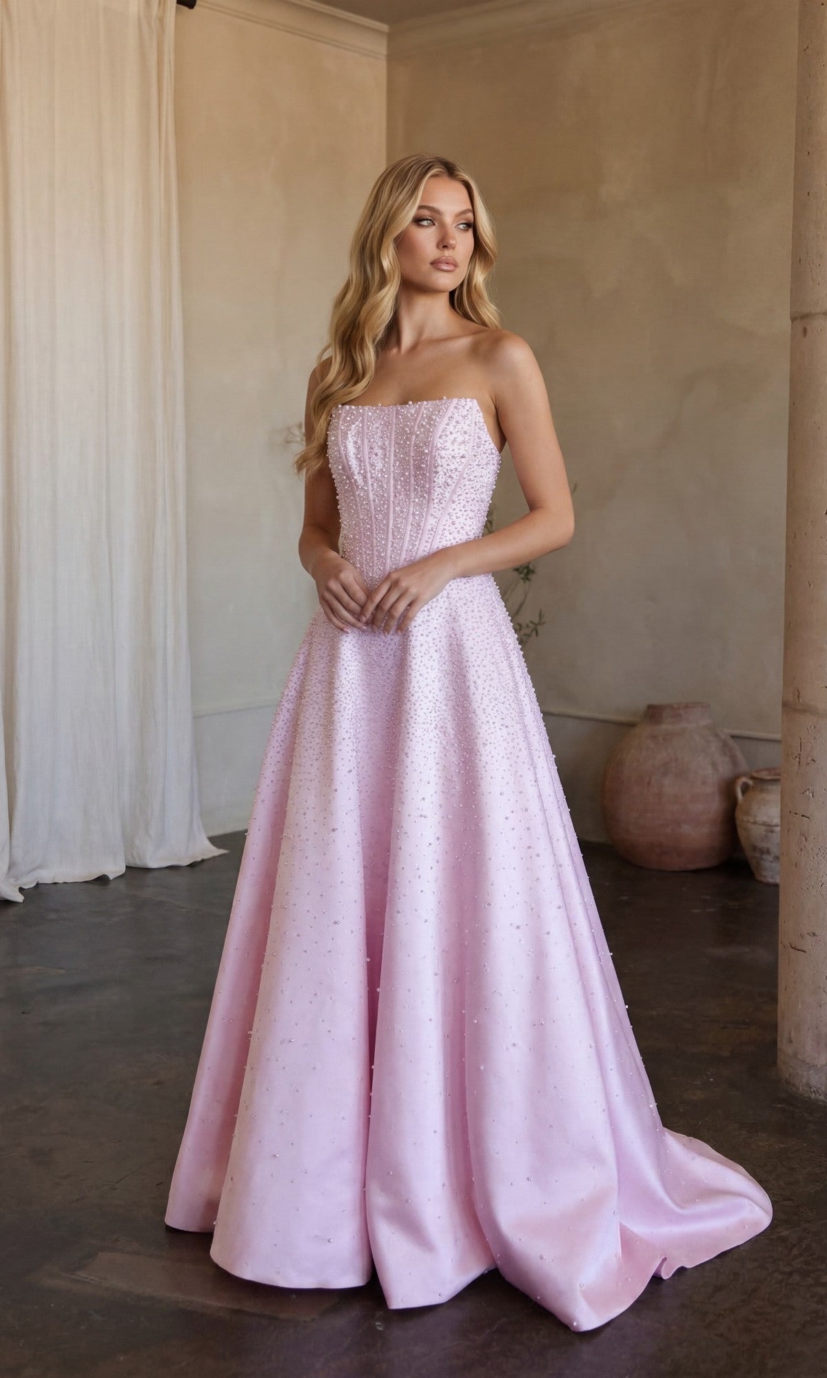 A woman stands indoors wearing the Ava Presley 47283 Strapless Long Pearl Prom Dress in blush, featuring a fitted bodice and flared floor-length skirt with delicate embellishments.