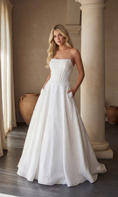A woman stands indoors wearing the Ava Presley 47283 Strapless Long Pearl Prom Dress in ivory, featuring a fitted bodice, full skirt with subtle beadwork, and pockets.