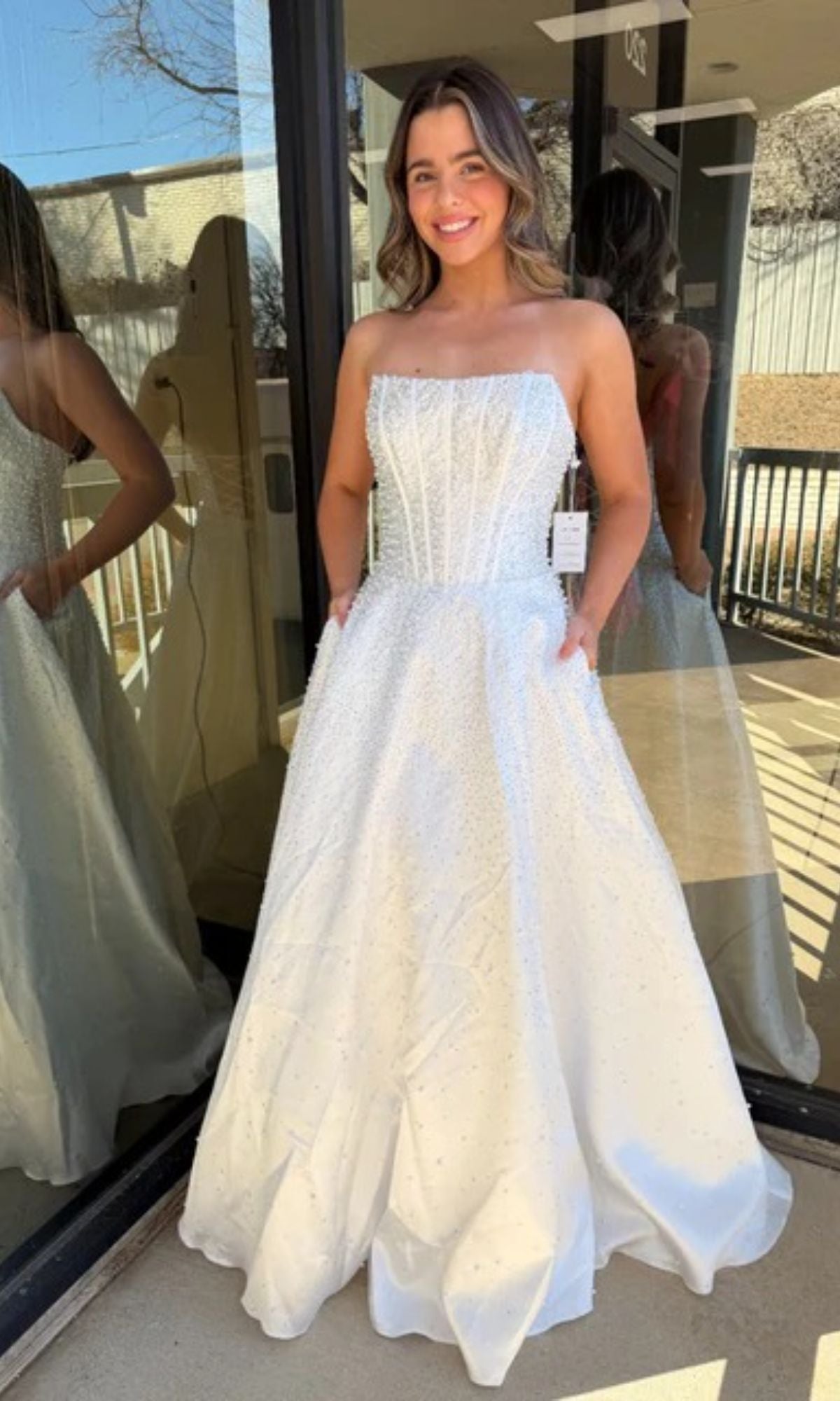 A young woman poses outdoors, smiling at the camera in the Ava Presley 47283 long prom dress, featuring a strapless fitted corset bodice, full skirt, and pockets.