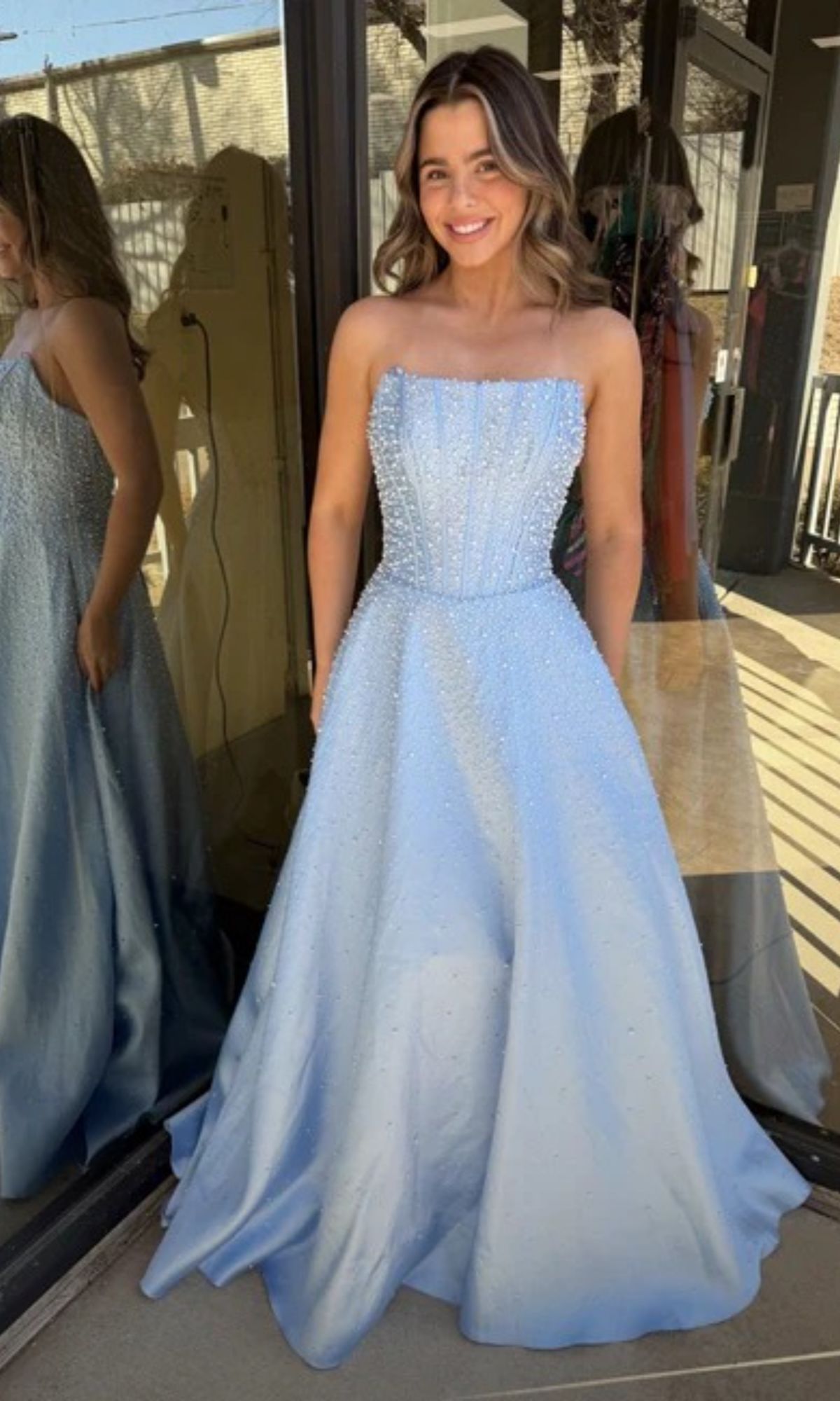 A young woman smiles at the camera outside a building, wearing the Ava Presley 47283 long prom dress—featuring a strapless corset bodice and beaded detailing.