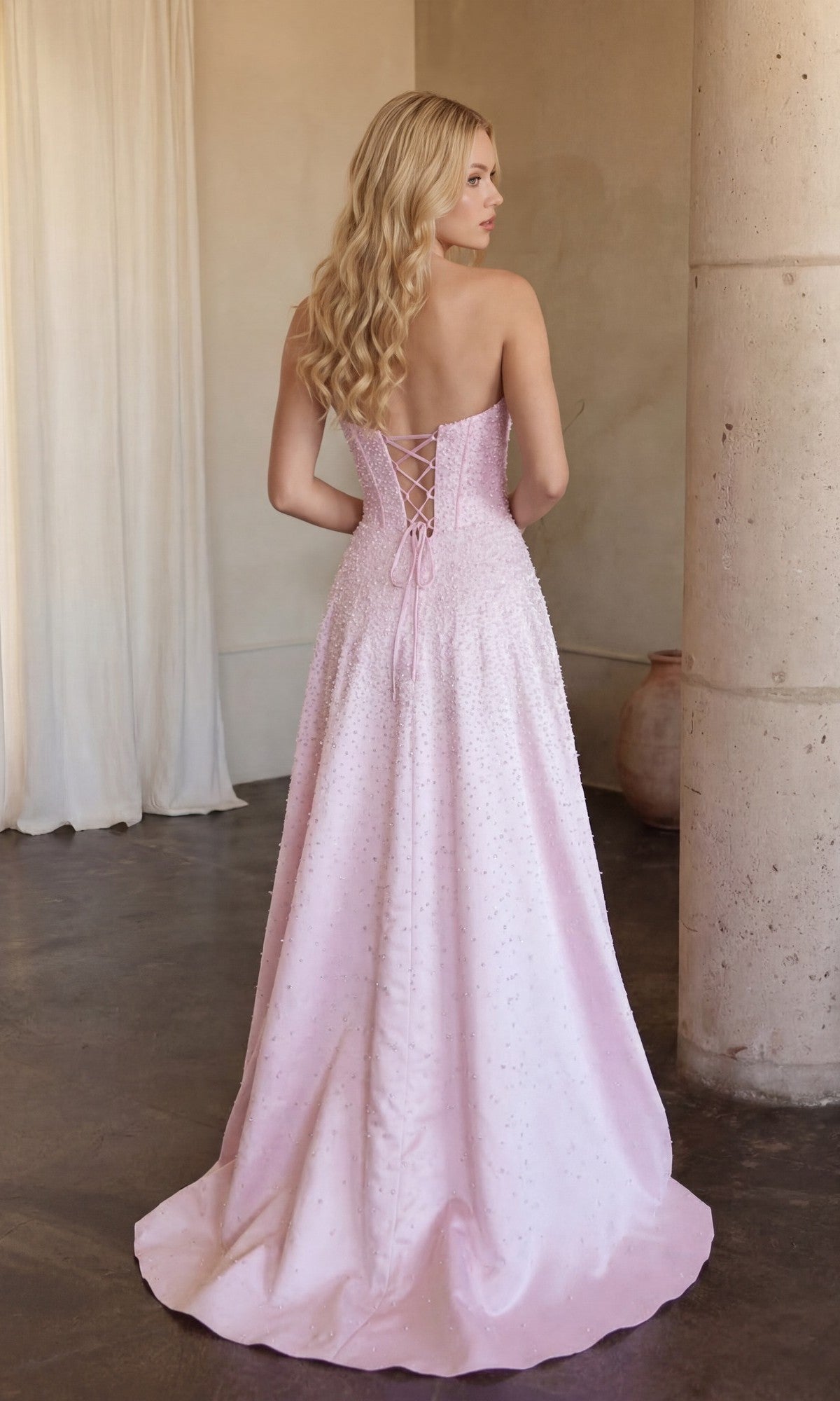 A woman with long blonde hair wears the Ava Presley 47283 Strapless Long Pearl Prom Dress, featuring a lace-up back. She stands indoors beside a pillar and white curtains.