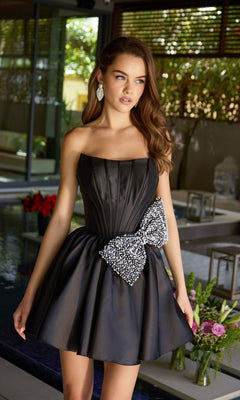 A woman in a Short Homecoming Dress: Ava Presley 47298 in black, featuring a strapless design with a boned corset bodice and large decorative bow, stands indoors near a pool with floral arrangements.
