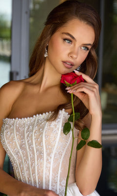 A woman stands indoors by a window, holding a red rose near her face and wearing the Ava Presley 47306 short cocktail dress adorned with pearls.