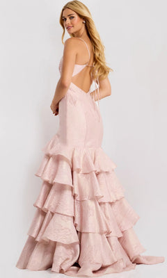 A woman models the Jovani 47329 long prom dress in pale pink, featuring an open back and tiered ruffle skirt, posing slightly away to showcase the gown’s elegant details against a plain background.
