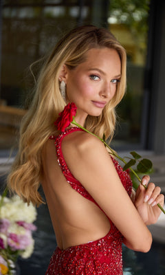 A woman with long blond hair and drop earrings stands indoors near a pool and vibrant flowers, holding a red rose over her shoulder while wearing the open-back "Ava Presley 47334" short cocktail dress.