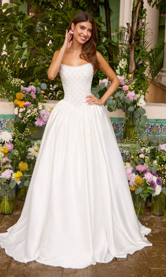 A woman wears an Ava Presley 47364 strapless, floor-length white gown with a beaded bodice and a full, smooth skirt, standing in front of floral arrangements.
