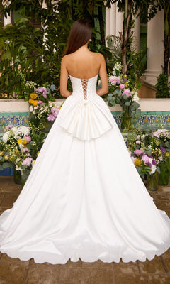 Back view of a white Ava Presley 47364 ball gown with a strapless, floor-length lace-up bodice and a large waist bow, standing before assorted flower arrangements.