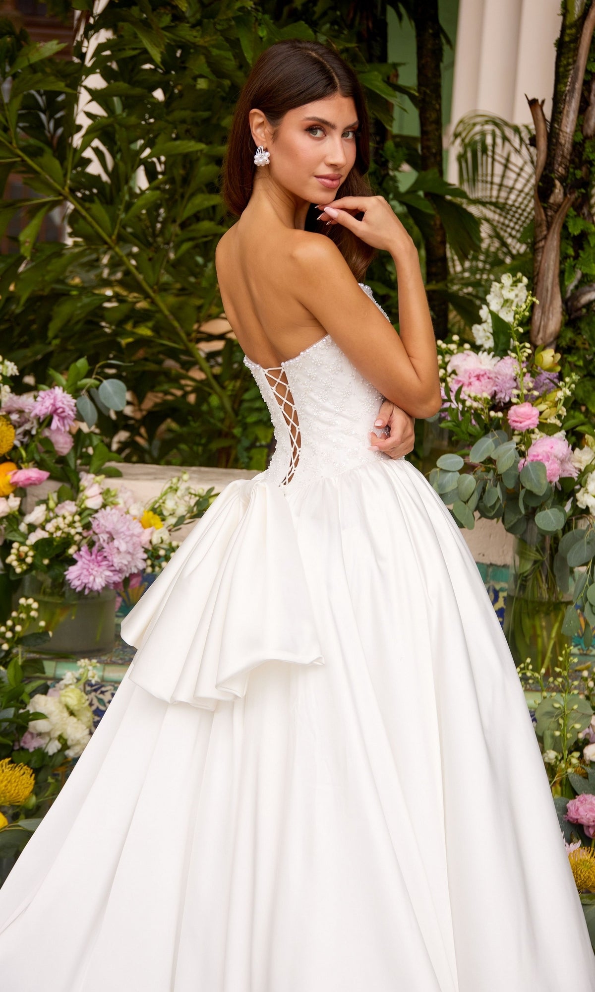 A person wearing a white, floor-length Ava Presley 47364 strapless ball gown with a lace-up back and a bow at the waist, standing in front of floral arrangements.