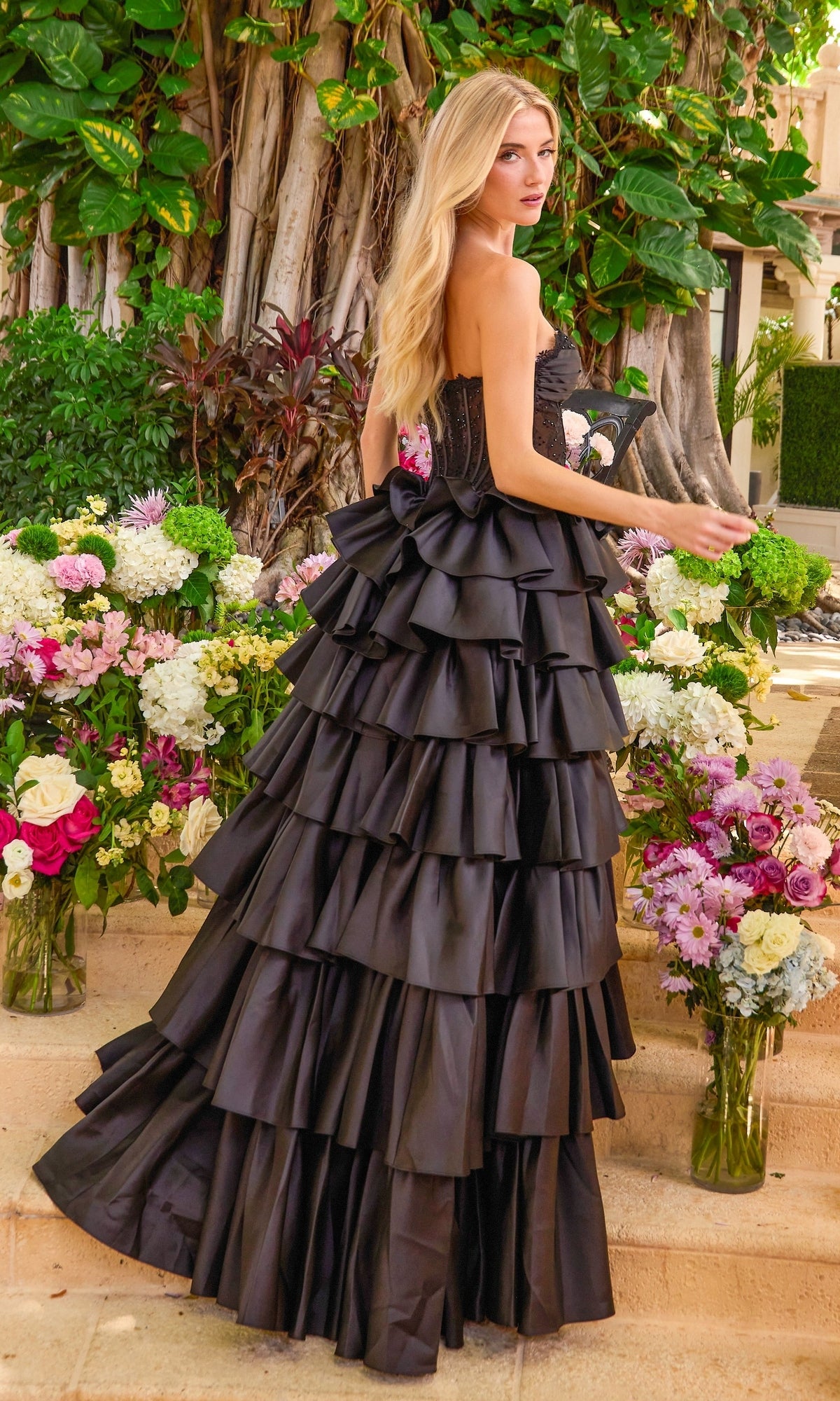 A woman wearing a strapless black corset Long Prom Dress: Ava Presley 47372 with a ruffled, tiered skirt, standing among floral arrangements.