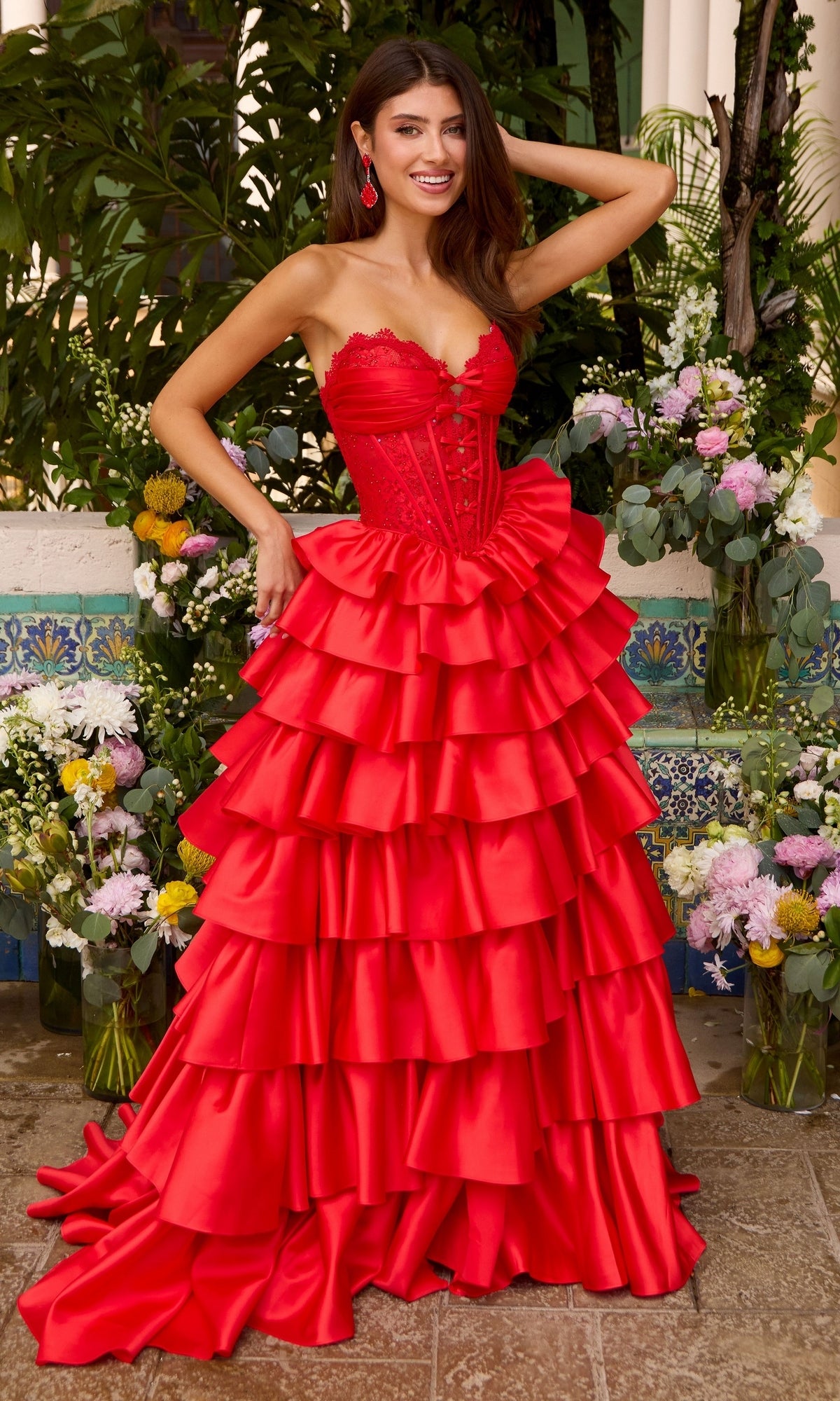 A model wears a red, strapless, floor-length Ava Presley 47372 corset prom dress with a tiered, ruffled skirt, standing in front of floral arrangements.