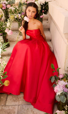 A person wears a minimalist, floor-length red Long Prom Dress: Ava Presley 47374 while sitting on stone steps and holding a pink flower.