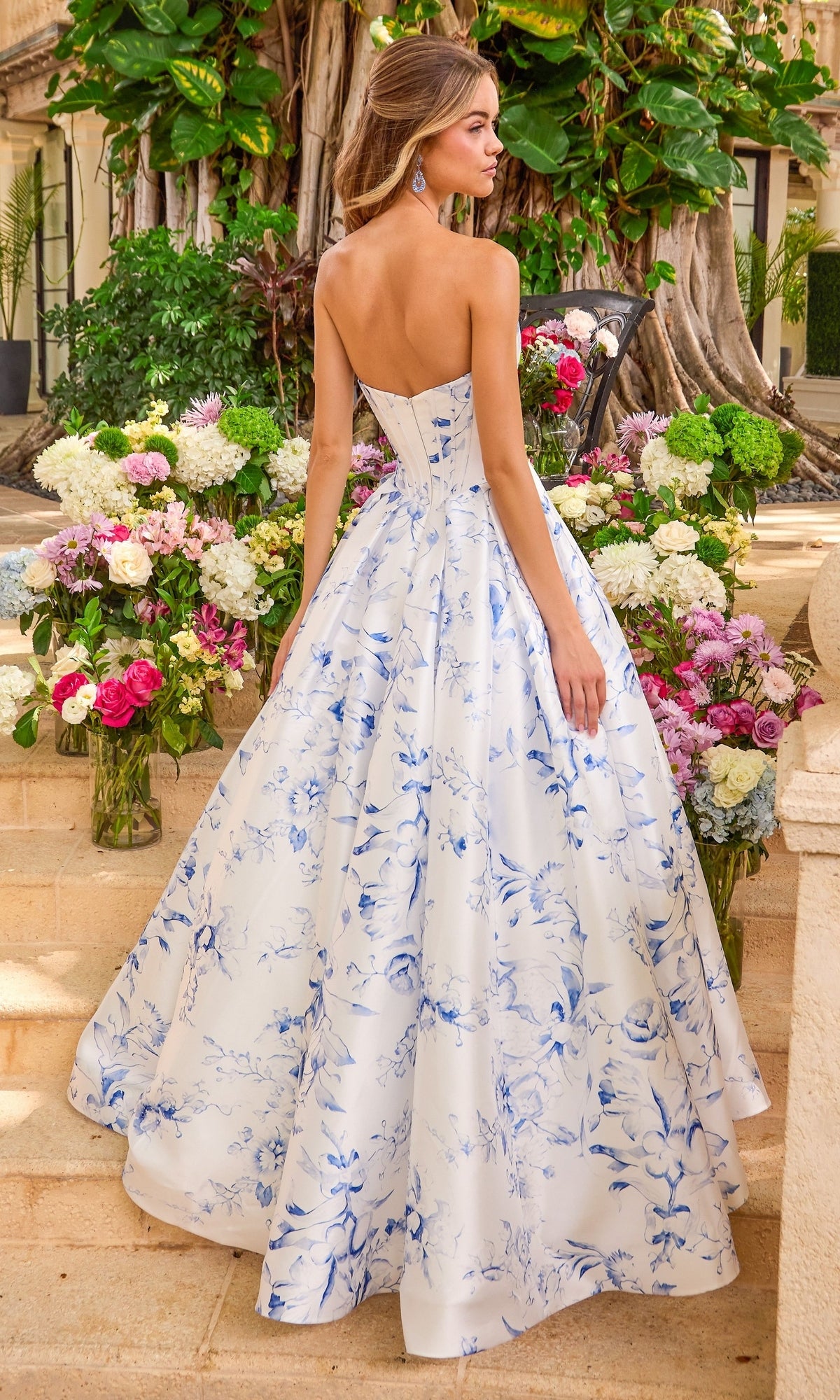 A woman wearing a Long Prom Dress: Ava Presley 47377 with a blue and white floral print, standing on stone steps surrounded by vases of flowers.
