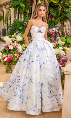 A woman wears a strapless, white and blue floral Ava Presley 47377 floor-length prom dress with a bodice cutout, posed outdoors among vases of flowers.