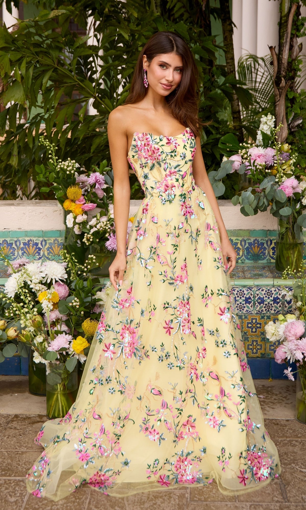 Woman wearing a strapless, floor-length Ava Presley 47391 yellow prom dress with floral embroidery, standing in front of flower arrangements.
