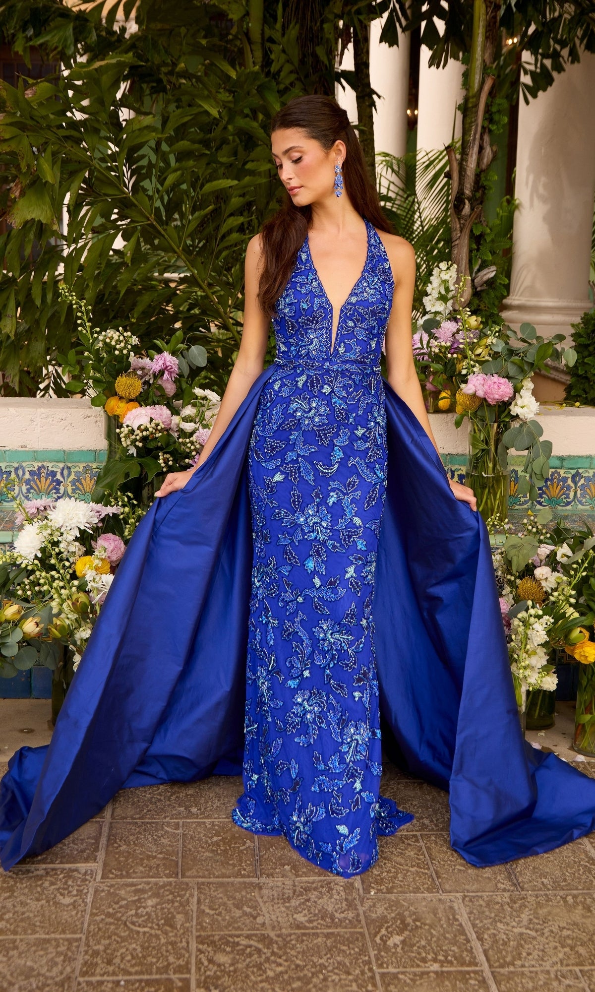 A woman wears an Ava Presley 47421 blue beaded halter prom dress featuring a floor-length silhouette, a plunging neckline, and a detachable overskirt.