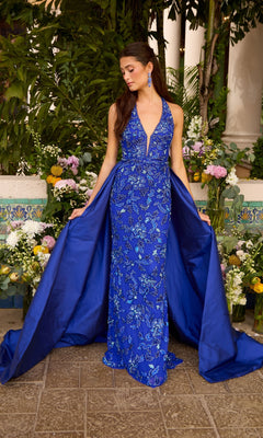 A woman wears an Ava Presley 47421 blue beaded halter prom dress featuring a floor-length silhouette, a plunging neckline, and a detachable overskirt.