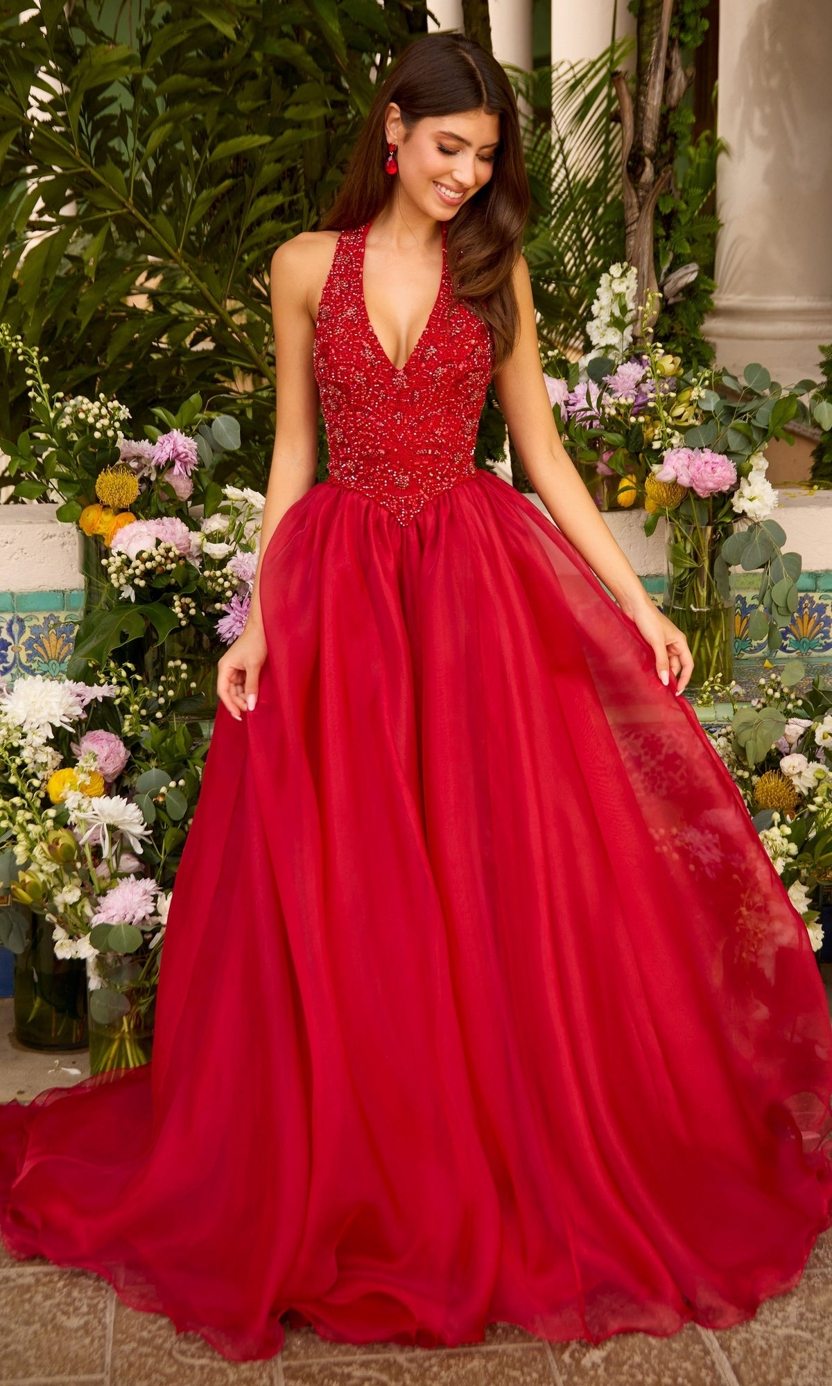 A woman wears a red Long Prom Dress: Ava Presley 47425, a beaded ball gown featuring a V-neck bodice and a full, layered, floor-length skirt.