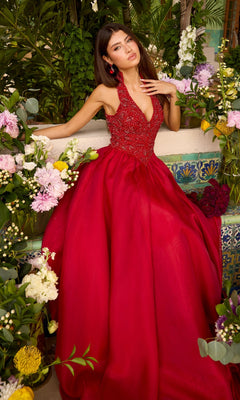 A person wearing a floor-length red Ava Presley 47425 beaded ball gown with a full skirt, seated among various floral arrangements.