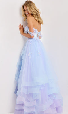 A woman wearing the Jovani 47427 long prom dress, a strapless pastel blue and lavender lilac ballgown with floral appliqués, stands against a white wall, smiling and turning slightly to the side.