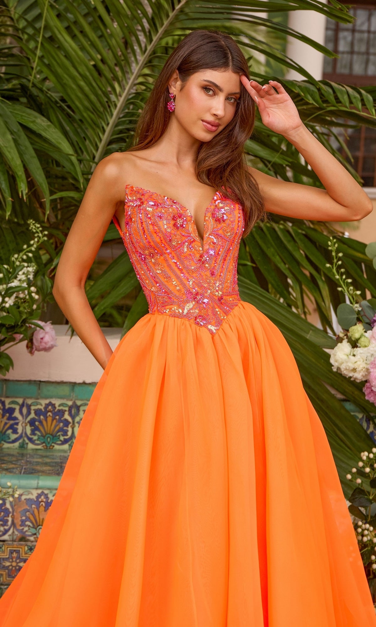 A person wears an Ava Presley 47430 strapless, neon orange ball gown with a sequined, pink-accented bodice and a full, floor-length skirt.