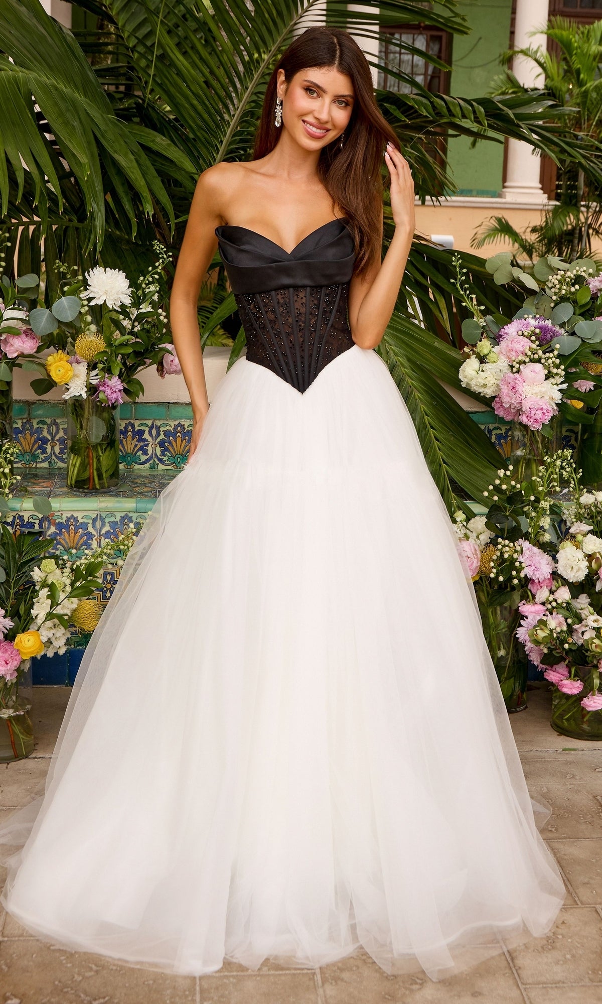 A woman wears an Ava Presley 47484 strapless corset ball gown with a black bodice and a full white tulle skirt, standing in front of floral arrangements.