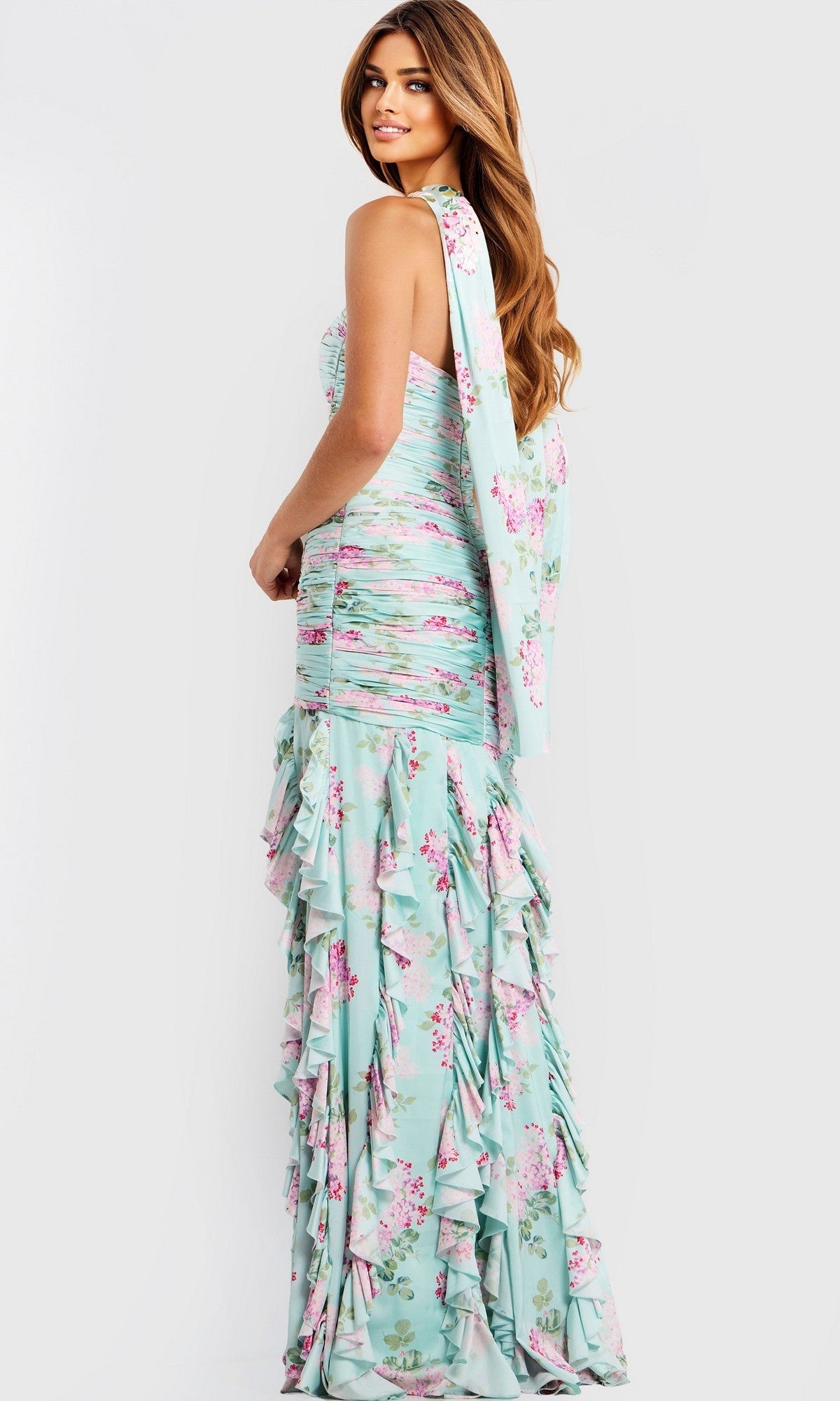 A woman stands sideways against a plain background, wearing the Jovani 47580 Long Prom Dress—a fitted, strapless floral chiffon gown with ruffled layers and a long sash.