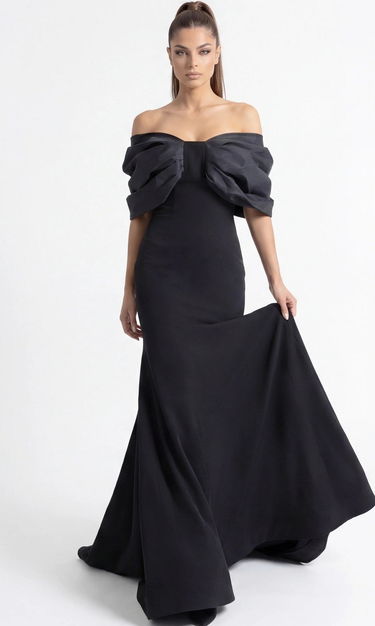 A woman wearing the Long Formal Dress: Jovani 47657—a fitted, off-the-shoulder black designer gown featuring oversized bow detailing on the bodice—stands against a plain white background, holding the skirt with one hand.
