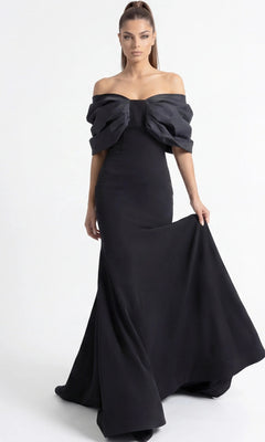 A woman wearing the Long Formal Dress: Jovani 47657—a fitted, off-the-shoulder black designer gown featuring oversized bow detailing on the bodice—stands against a plain white background, holding the skirt with one hand.