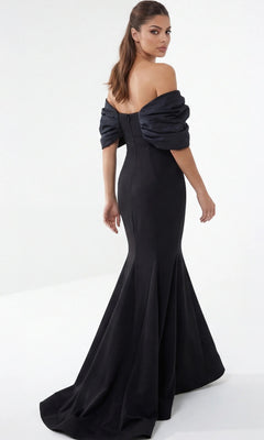 Woman in an off-the-shoulder, floor-length black Jovani 47657 long formal dress, standing and looking over her shoulder against a plain white background—a sophisticated choice for a designer gown or a modern mother-of-the-bride ensemble.