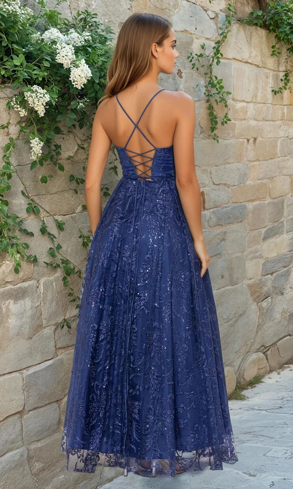 A woman in the Long Navy Formal Dress: Blondie Nites 4768BN with sequins, crisscross straps, and a lace-up back stands by a stone wall covered in green vines and white flowers.