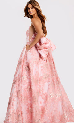 A woman looks over her shoulder in the Jovani 47702 long prom dress, a sleeveless pink gown with a floral pattern and a large bow at the back, set against a plain light background.
