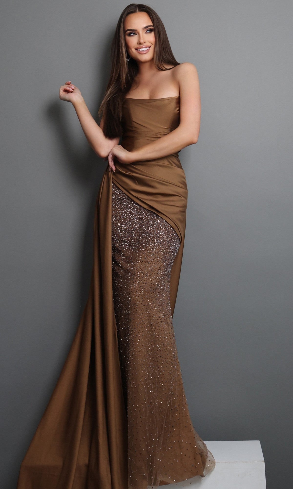 A woman stands against a gray background wearing the Jovani 48142 long formal dress in brown, featuring a strapless design with a sheer, beaded sheath skirt and an elegant draped satin overlay.