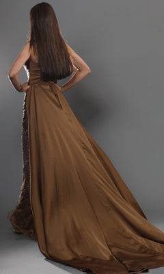 A person with long brown hair stands facing away, wearing the Jovani 48142 long formal dress—a strapless gown with a flowing train, detailed embellishments, and a draped satin overlay that adds depth and texture to the look—set against a neutral gray background.