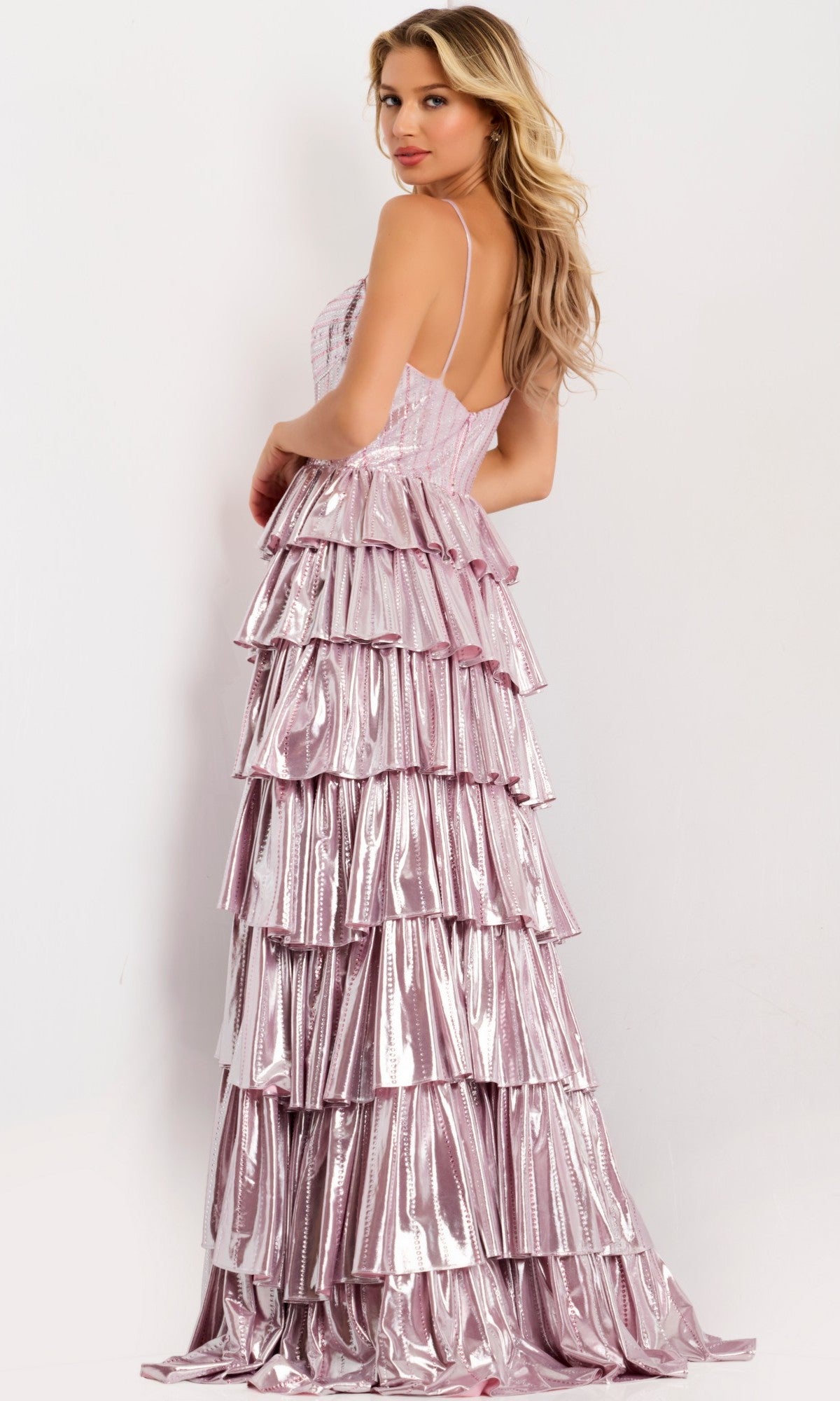 A woman stands sideways, looking over her shoulder, wearing the Jovani 48341 long prom dress—sleeveless and metallic pink with tiered ruffles—against a plain background.