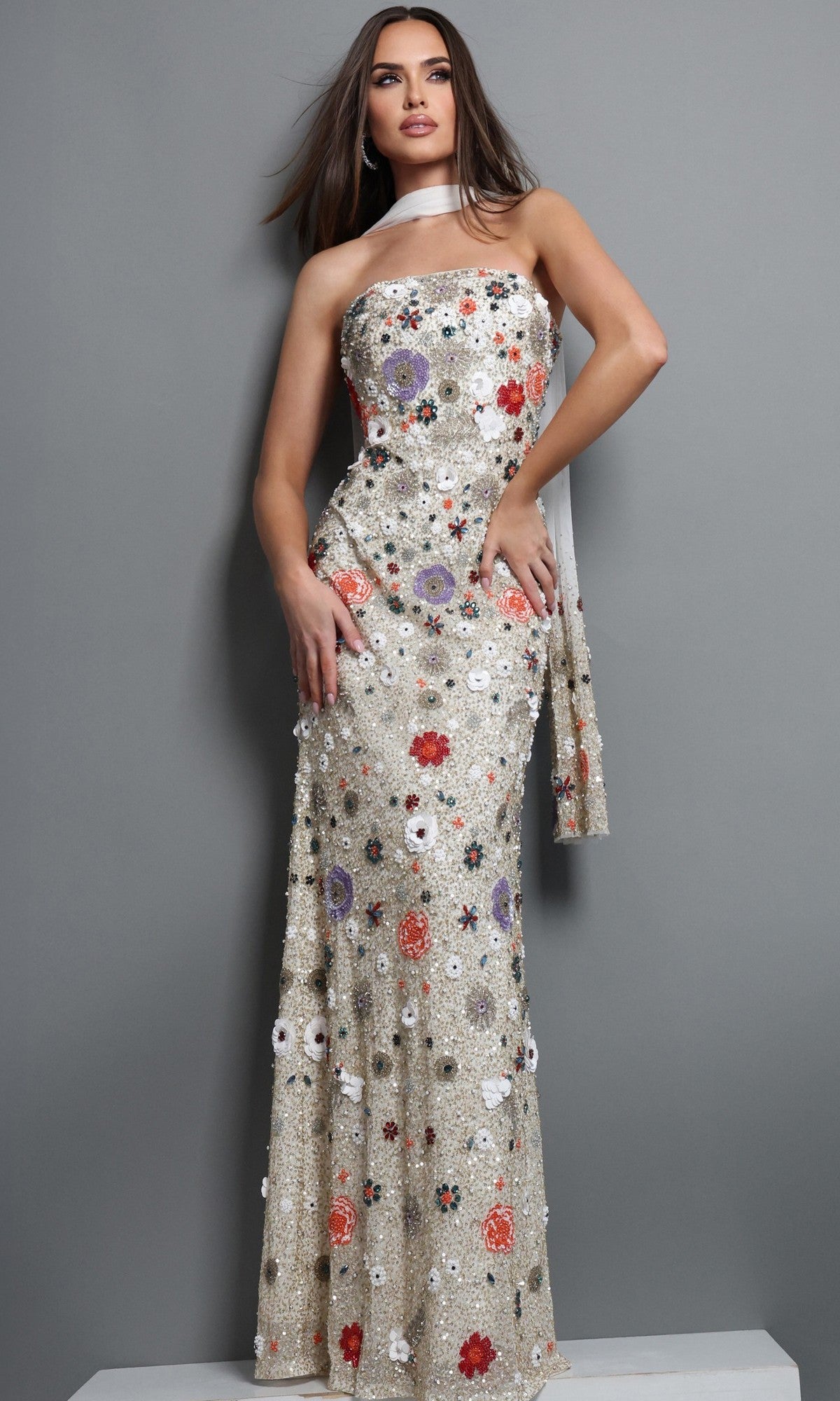 A woman stands against a gray background wearing the Long Formal Dress: Jovani 48587 in Ivory/Multi—an exquisite strapless, floor-length gown adorned with stunning red, purple, and white floral embroidery. A matching scarf is elegantly draped over her shoulder, perfectly completing this elegant look.