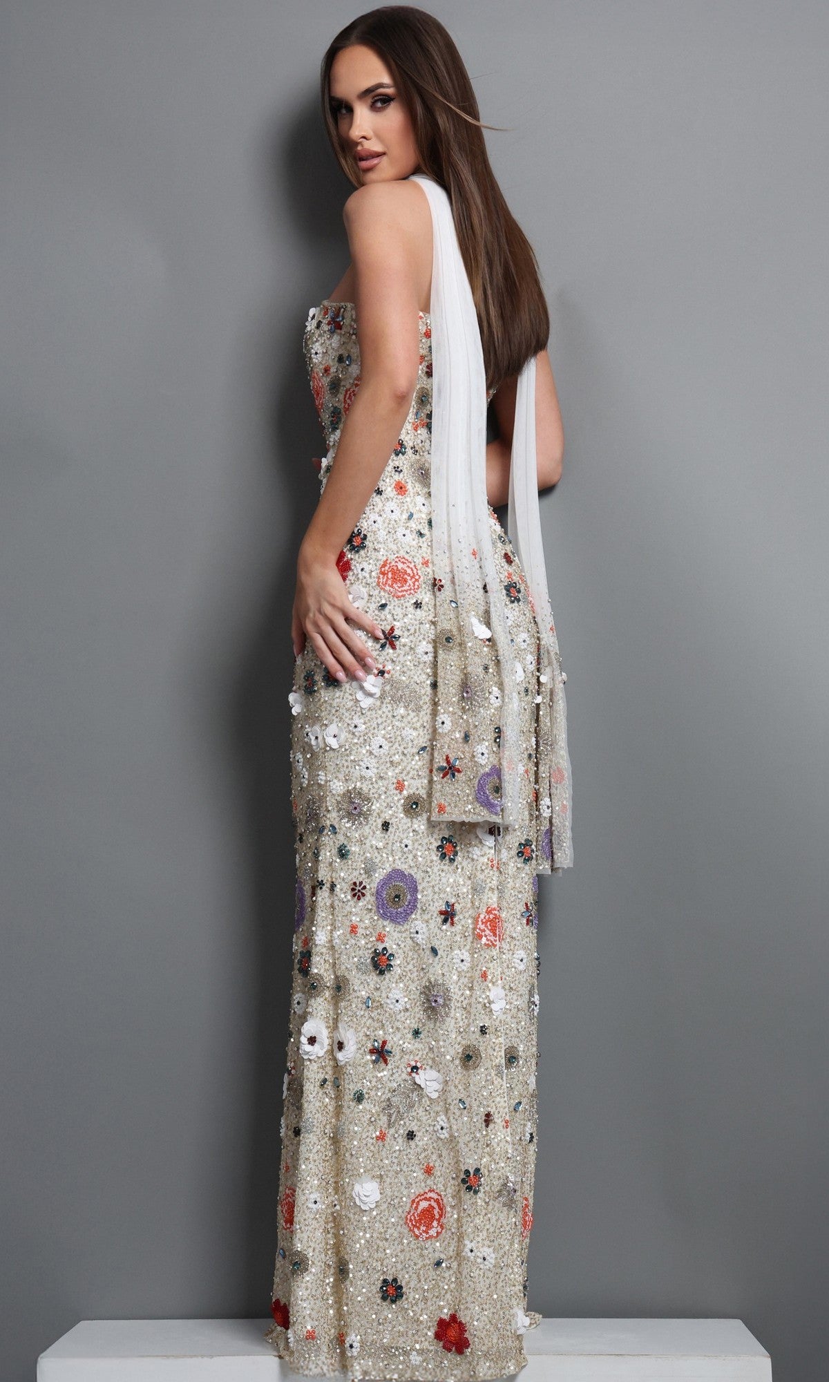 A woman wearing the Jovani 48587 long formal dress—a strapless ivory gown adorned with exquisite floral embroidery—stands on a platform, positioned sideways against a plain gray background.