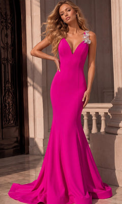 A woman stands in front of a stone building wearing the Jovani 48663 long formal dress in fuchsia—a floor-length, sleeveless gown with a deep V neckline, floral embellishment on one shoulder, and an elegant mermaid hem.