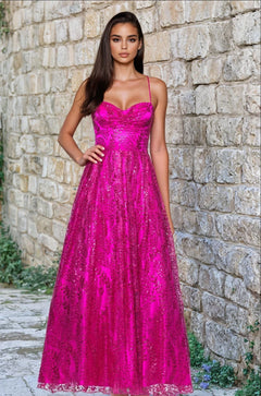 A woman stands outdoors by a stone wall, wearing the Blondie Nites 4867BN Long Glitter Prom Dress in hot pink, a junior-size formal gown with thin straps and floor-length sequin embellishments.