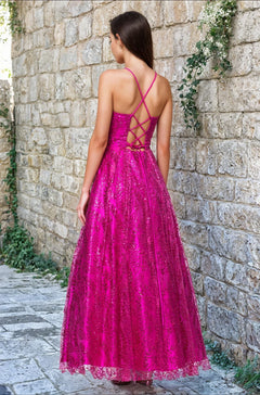 A woman stands on a stone path facing a stone wall, wearing the Blondie Nites 4867BN Long Glitter Prom Dress—a bright pink, floor-length junior formal dress with a crisscross lace-up back and shimmering glitter-print details.