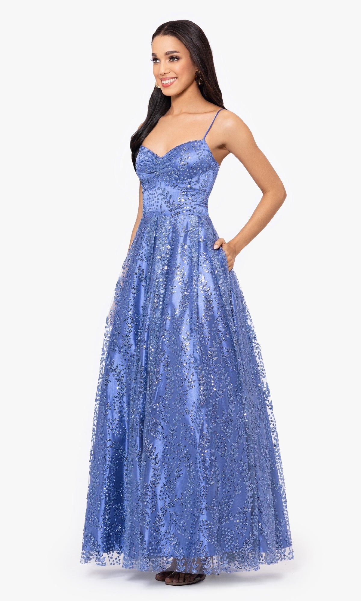 A woman wears the affordable Blondie Nites 4867BN, a long blue glitter prom dress with thin straps and a full skirt, standing against a plain light background.