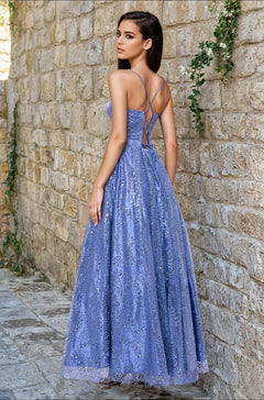 A woman in the affordable Blondie Nites 4867BN long glitter prom dress—a sparkly lavender gown with crisscross straps—poses outdoors against a stone wall.